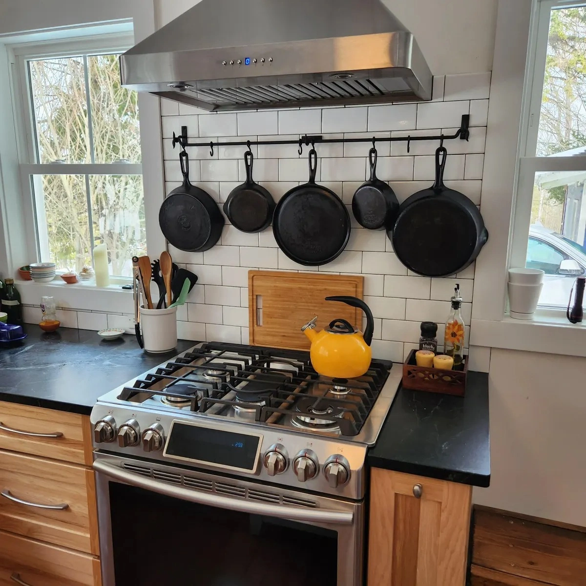 Modern kitchen with black cast iron pans hanging on a wall-mounted pot rack above a stainless steel gas stove with yellow kettle